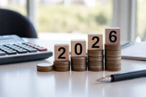 Holzwürfel mit der Jahreszahl 2026 auf ansteigenden Münzstapeln neben Taschenrechner und Kugelschreiber